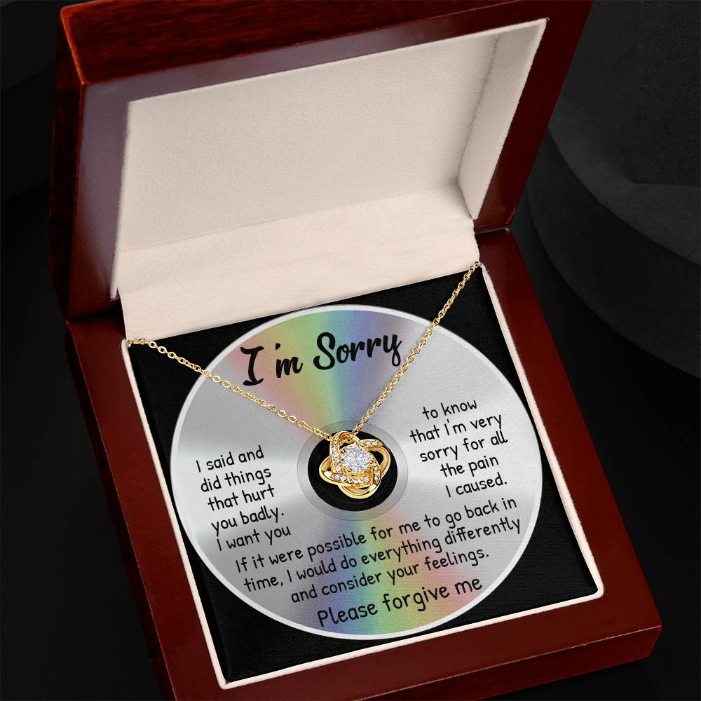 The Sorry-Back In Time - Love Knot Necklace with a yellow gold finish rests on a CD inside an open wooden box. The pendant features stunning cubic zirconia crystals. The CD displays an apology message: "I'm Sorry" at the top and "Please forgive me" at the bottom.