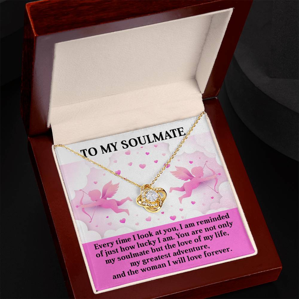 The Soulmate-Greatest Adventure Love Knot Necklace, with a gold finish, rests in a jewelry box labeled "To My Soulmate." It is set against pink Cupid illustrations and a heartfelt message, making it an ideal gift.