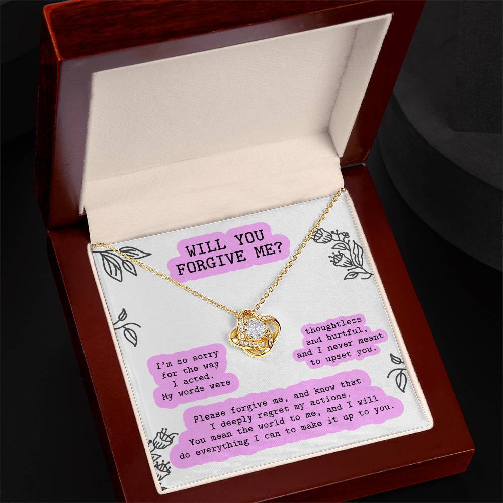 The "Sorry-Never Meant To - Love Knot Necklace," adorned with cubic zirconia crystals, rests on a cushion in an open box. The necklace, crafted with two interlocking hearts in a yellow gold finish, is accompanied by a heartfelt message asking for forgiveness and expressing regret for hurtful words.