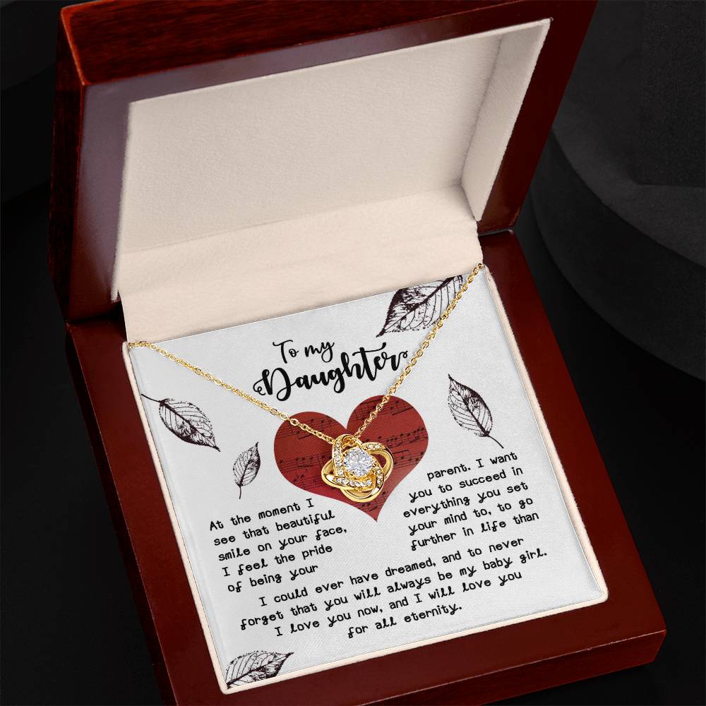 The "Daughter-On Your Face - Love Knot Necklace" features an intertwined heart pendant adorned with cubic zirconia crystals, presented in an open box. The lid of the box displays a heartfelt message for a daughter, embellished with decorative elements. This stunning necklace is available in both white gold and yellow gold finishes.