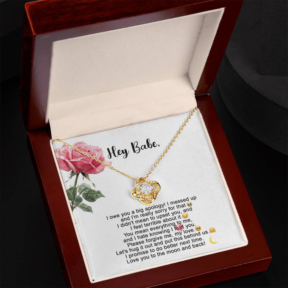 The "Sorry-Hug It Out - Love Knot Necklace" features an 18k yellow gold pendant adorned with small cubic zirconia crystals. It comes on a card decorated with roses, accompanied by the message "Hey Babe" and an apology note.