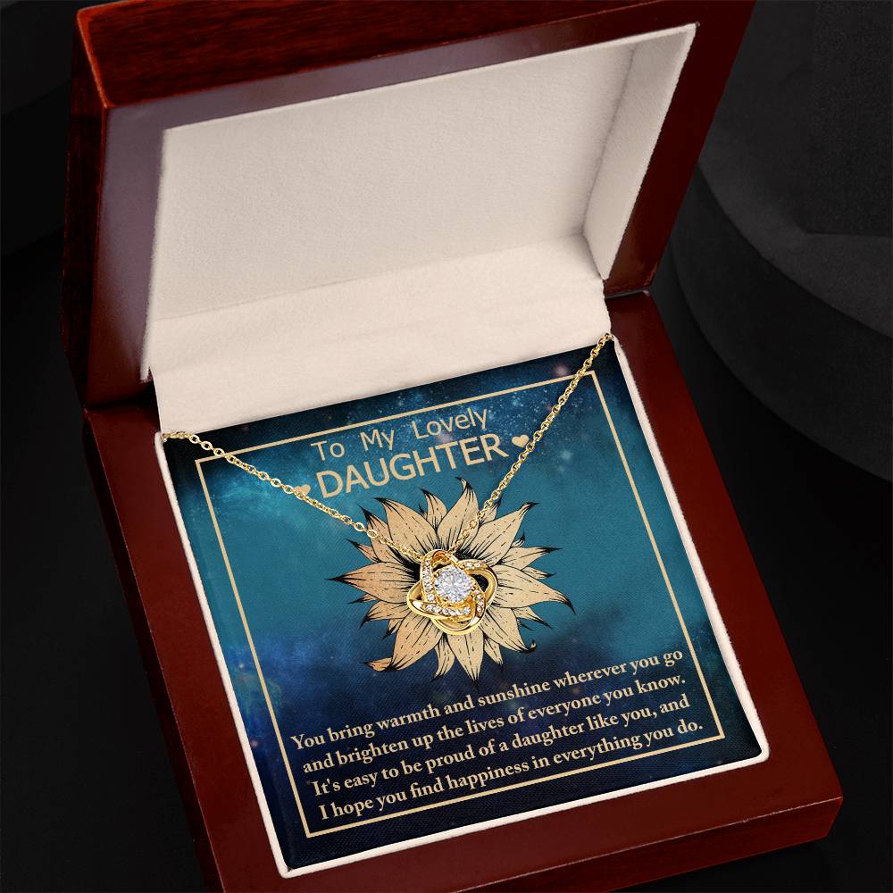 The "To Daughter, Be Proud Of - Love Knot Necklace" is a gold piece showcasing a sunflower pendant with a cubic zirconia gem at its center. It is elegantly presented in an open wooden box lined with white fabric. The inside of the lid features a card that reads, "To My Lovely Daughter," accompanied by a heartfelt message in white text on a blue background.