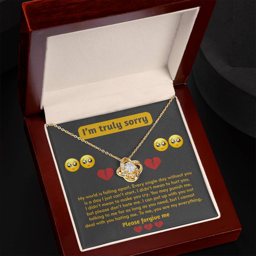 Nestled inside an open wooden box, a gold-finished heart-shaped necklace with a cross pendant is accompanied by the "Sorry-Make You Cry - Love Knot Necklace" shimmering in cubic zirconia. Above an apology note featuring crying emoji graphics on the inside lid, the inscription "I'm truly sorry" can be seen.