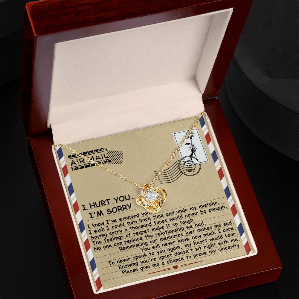 The "Sorry-Makes Me Sad - Love Knot Necklace," featuring a gold interwoven heart pendant embellished with cubic zirconia crystals, is beautifully presented in an open wooden box alongside an apology card printed in black text on a vintage airmail envelope design.