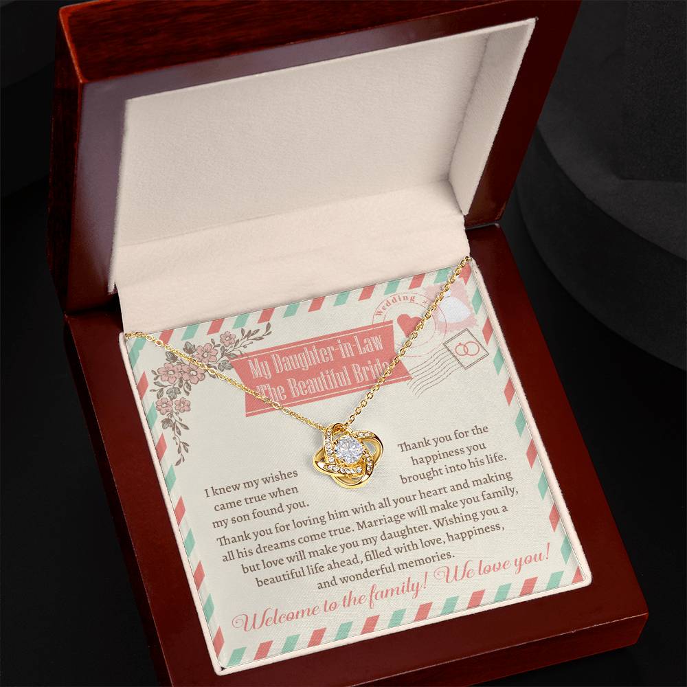 A "To My Daughter-In-Law, Make You Family - Love Knot Necklace" with an intertwined design is displayed on a heartfelt message card for a daughter-in-law, expressing love and welcoming her to the family. The piece, available in white gold or yellow gold finish, is adorned with shimmering cubic zirconia.