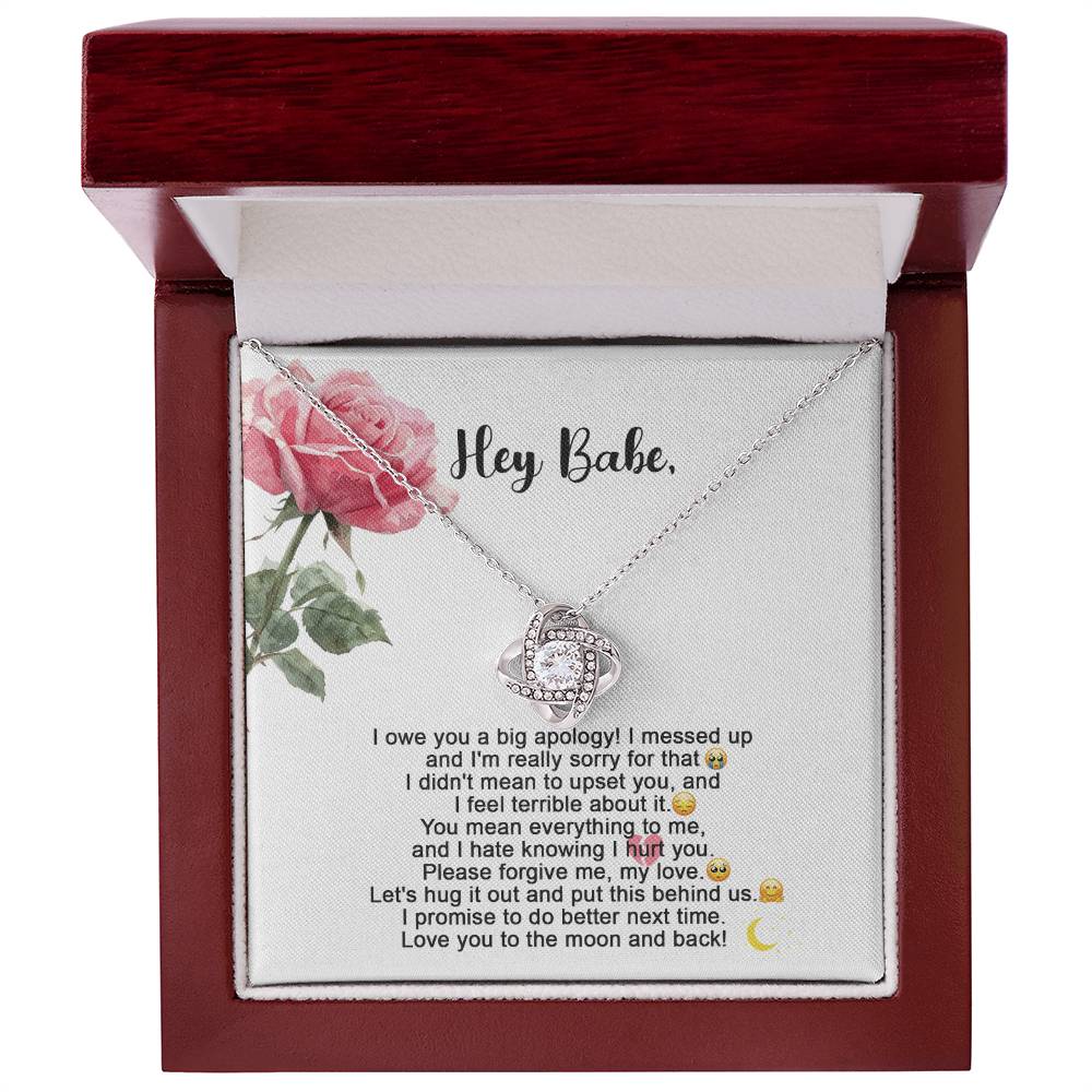 The "Sorry-Hug It Out - Love Knot Necklace" features an 18k yellow gold pendant adorned with small cubic zirconia crystals. It comes on a card decorated with roses, accompanied by the message "Hey Babe" and an apology note.