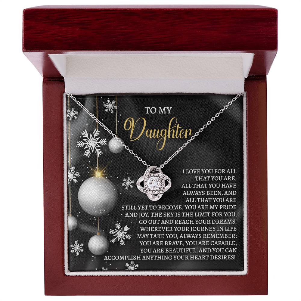 The Daughter-Pride And Joy - Love Knot Necklace is nestled in a box with a heartfelt message for a daughter, featuring a decorative background of snowflakes and ornaments. This personalized gift sparkles with cubic zirconia, making it a shimmering token of affection.