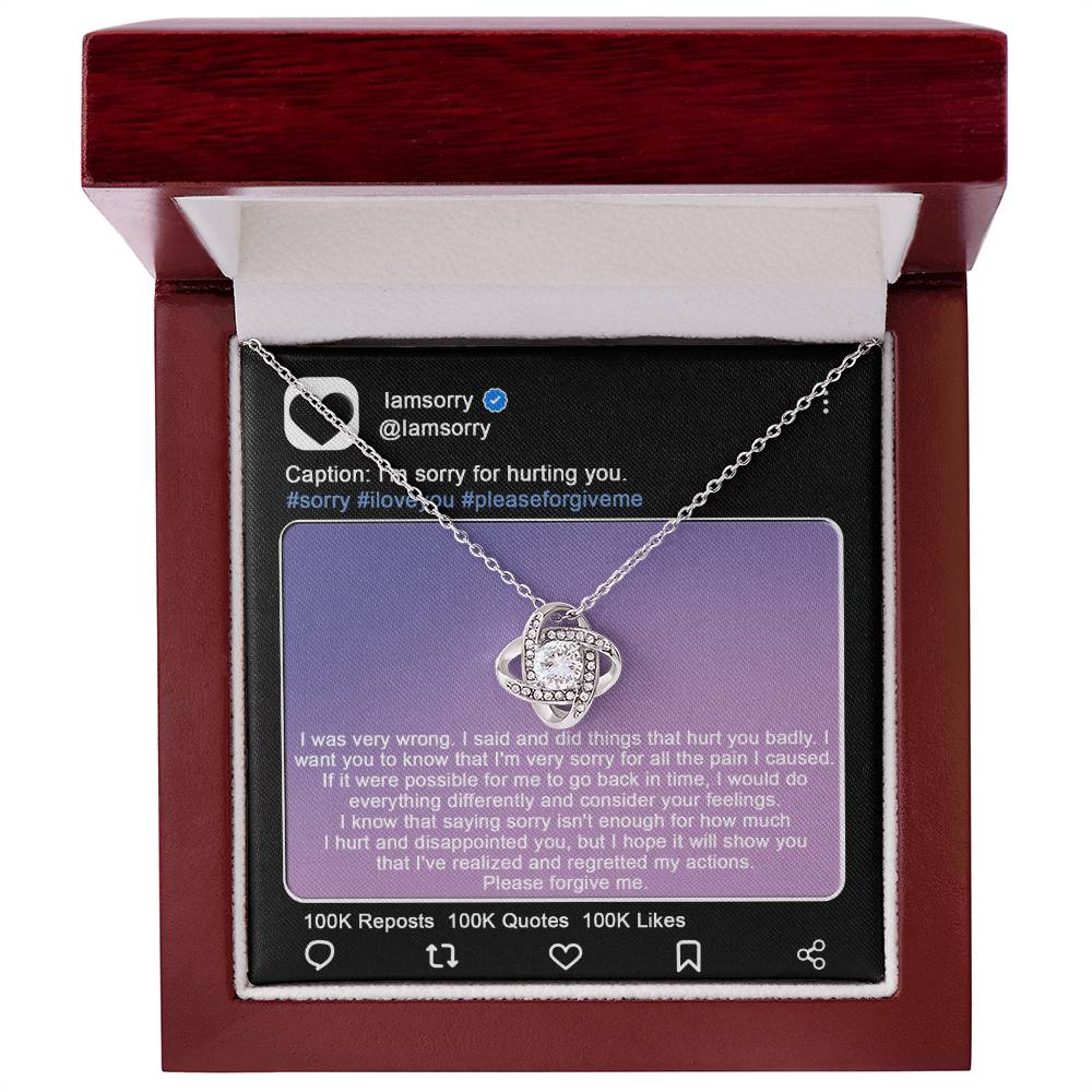 A stunning "Sorry-Hurt You Badly - Love Knot Necklace" in 14k white gold, adorned with cubic zirconia crystals, is displayed in front of a printed Instagram post that includes an apology message and hashtags like #sorry and #iloveyou.