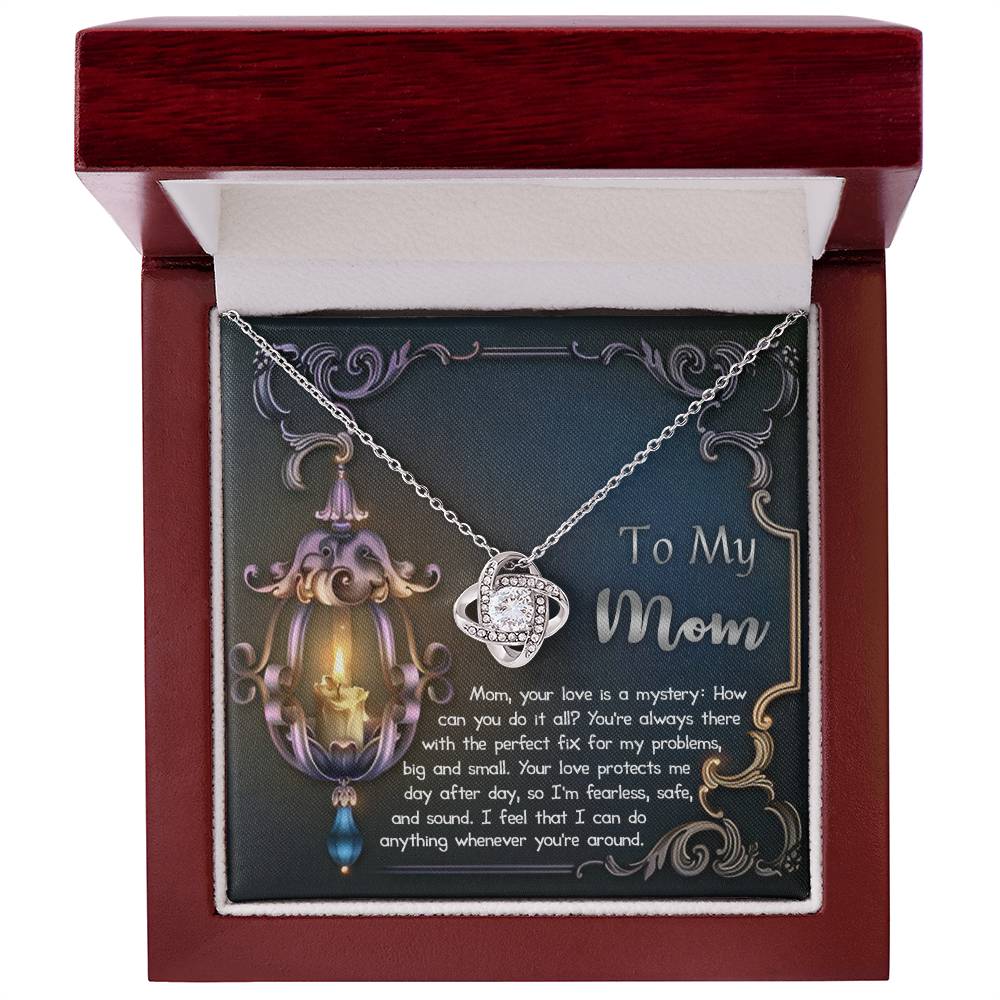 The "To Mom, Love Is Mystery - Love Knot Necklace" features a heart-shaped pendant with a gold finish and shimmering cubic zirconia. It is presented in a red jewelry box, with the interior lid displaying the heartfelt message "To My Mom.