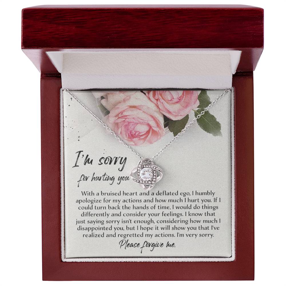 The "Sorry-Regretted My Actions - Love Knot Necklace," crafted in 14k white gold with an intricate pendant adorned with cubic zirconia crystals, is displayed against a backdrop featuring a printed apology note surrounded by roses. The note expresses regret and asks for forgiveness for past actions.