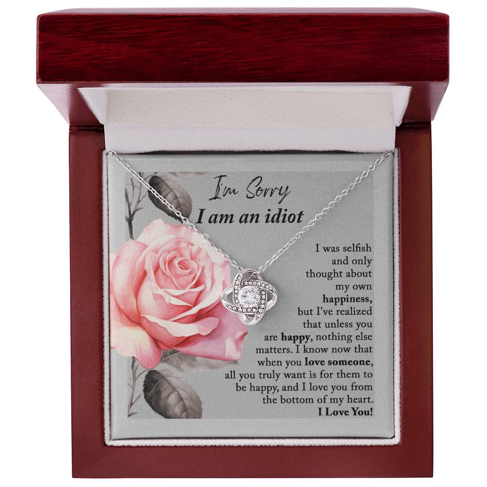 The "Sorry-Love Someone - Love Knot Necklace" is an elegant silver piece with a heart-shaped pendant, adorned with sparkling cubic zirconia crystals. It rests on a beautifully designed box featuring a pink rose and a heartfelt apology note, making it truly a classic gift.