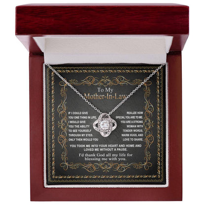 The "To Mother-In-Law, Through My Eyes - Love Knot Necklace" with a heart-shaped pendant rests in a maroon jewelry box. The inside of the lid features a personalized message addressed to a mother-in-law, expressing gratitude and appreciation. This elegant piece, adorned with premium cubic zirconia, makes for the perfect personalized gift.