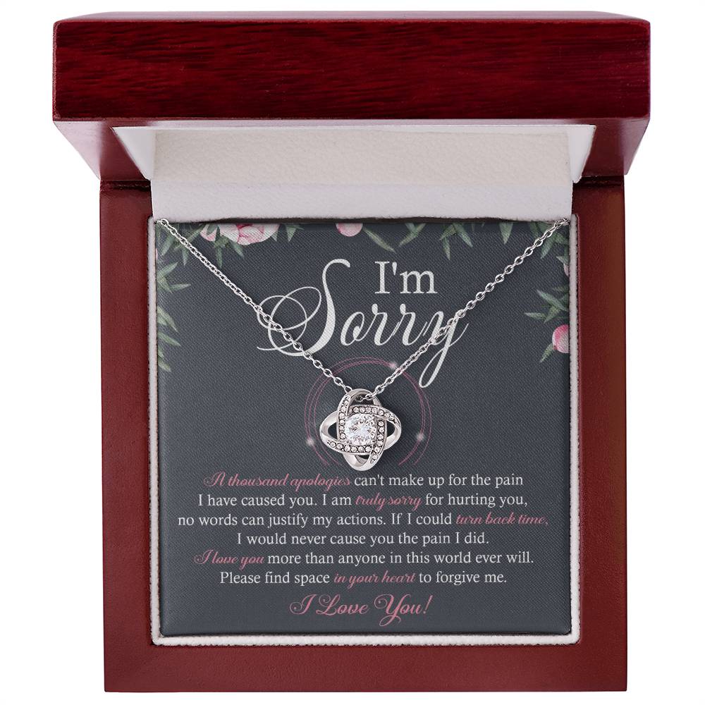 A box containing the "Sorry-World Ever Will - Love Knot Necklace," featuring a central pendant adorned with cubic zirconia crystals. The box lid displays text apologizing and seeking forgiveness, framed by a decorative floral border.