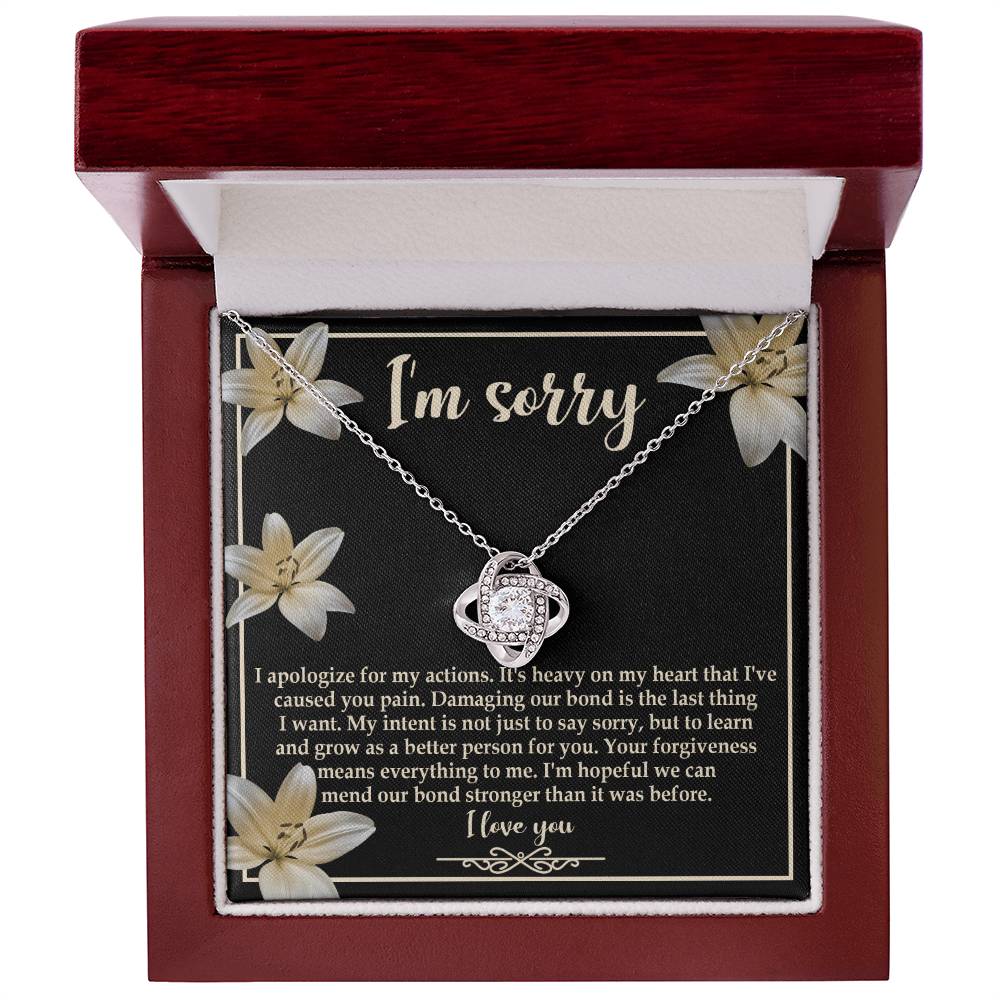 The "Sorry-The Last Thing - Love Knot Necklace" showcases a love knot pendant against a striking black background adorned with white flowers. It's accompanied by an apology message that reads, "I'm sorry. I apologize for my actions... I'm hopeful we can mend our bond stronger," and is beautifully finished with white gold and shimmering cubic zirconia crystals.