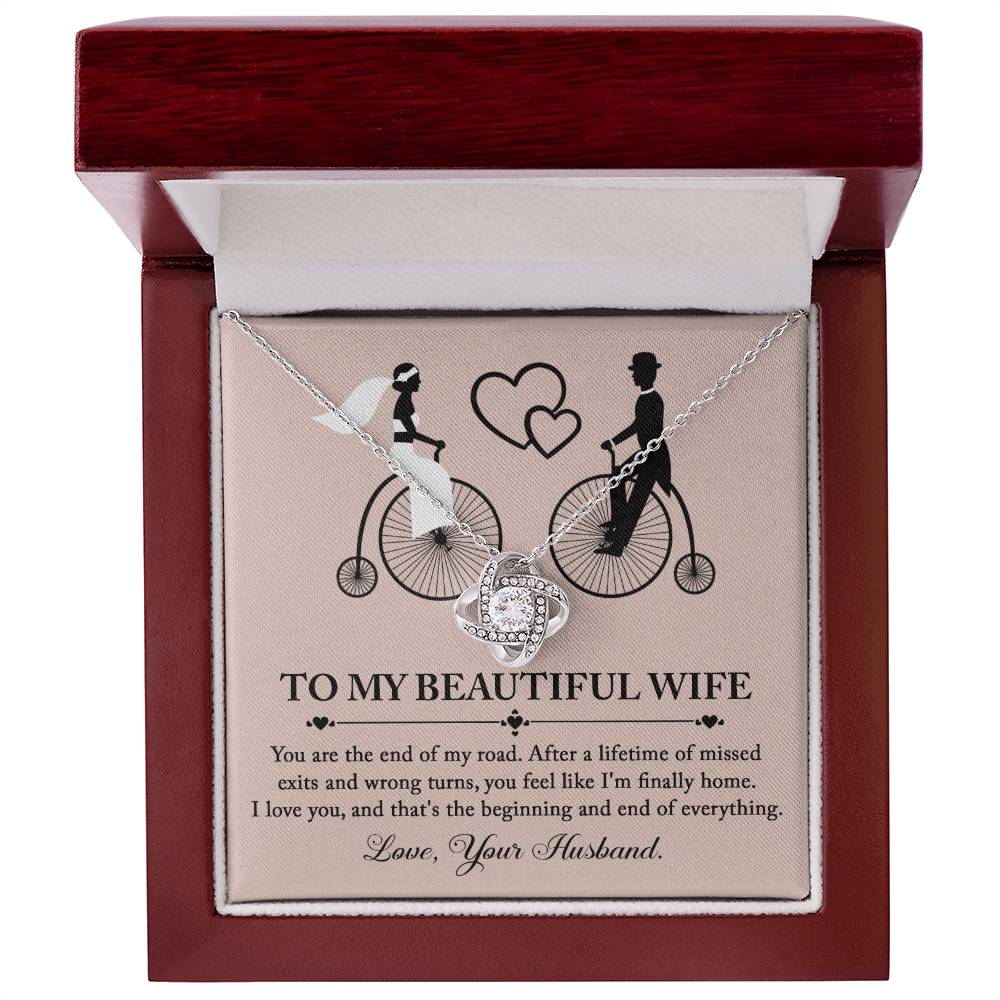 To Wife, I'm Finally Home - Love Knot Necklace with intertwined circle pendant on a card featuring an illustration of a couple on bicycles. The necklace, adorned with cubic zirconia crystals and a stunning gold finish, comes with a card that reads: 'To my beautiful wife' followed by a romantic message and ending with 'Love, Your Husband.'