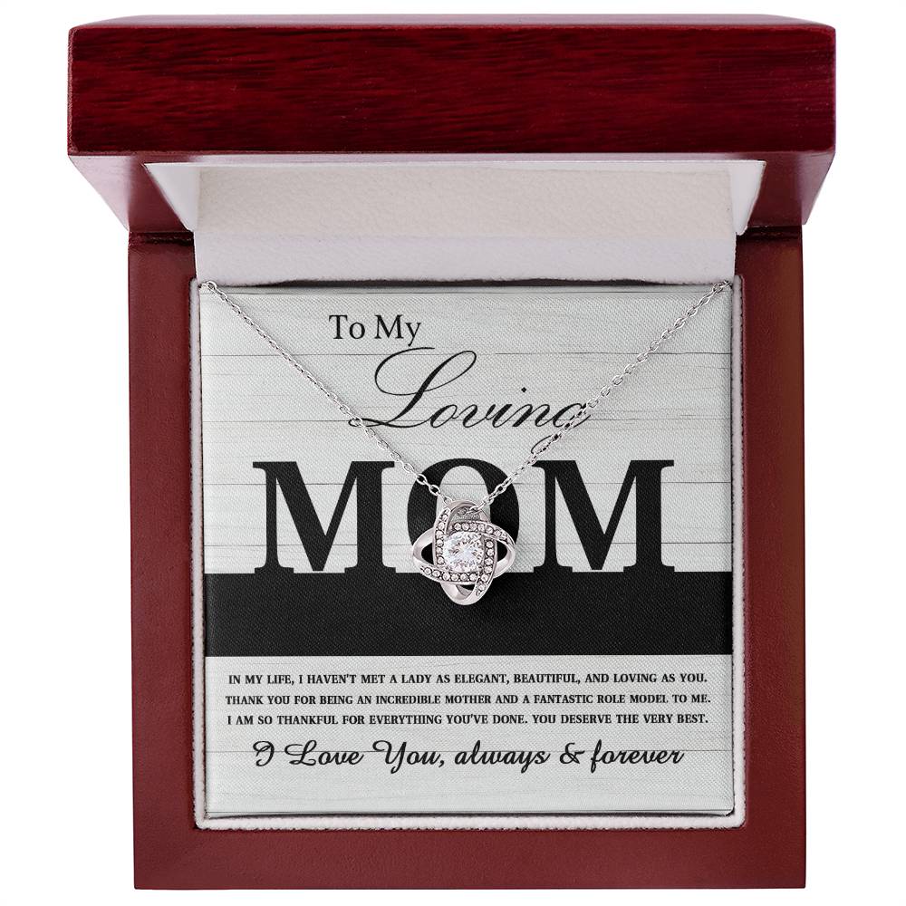 A "To Mom, Loving As You - Love Knot Necklace" adorned with shimmering cubic zirconia crystals sits elegantly in an open jewelry box. The lid of the box features an inscription expressing heartfelt love and gratitude for a mother, making it the perfect personalized gift.