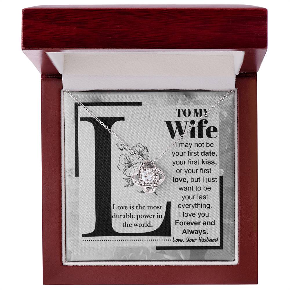 A "to Wife, Love Is - Love Knot Necklace" with a knot design is displayed on a card featuring a printed message to a wife about lasting love, signed from a husband. The piece sparkles with cubic zirconia crystals and boasts an elegant gold finish.