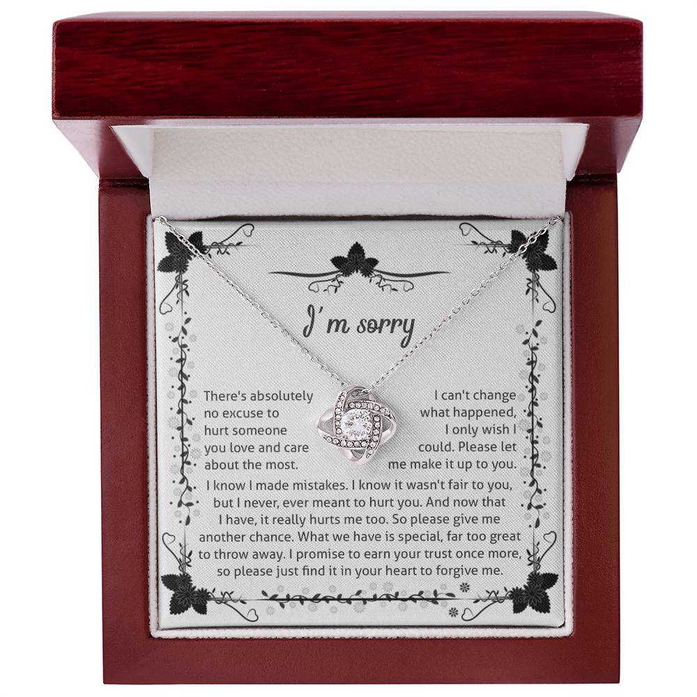 The Sorry-No Excuse - Love Knot Necklace, with a white gold finish, showcases a delicate knot design on a chain adorned with cubic zirconia crystals. The pendant is presented on a box featuring an "I'm sorry" message that expresses regret and seeks forgiveness, framed by a charming floral border at the top.