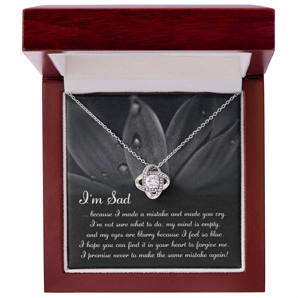 The "Sorry-Feel So Blue - Love Knot Necklace," featuring a stunning knot pendant adorned with cubic zirconia crystals, is displayed over a card with an apologetic message starting with "I'm Sad" and set against a floral background. This personalized gift beautifully combines elegance and heartfelt sentiment.