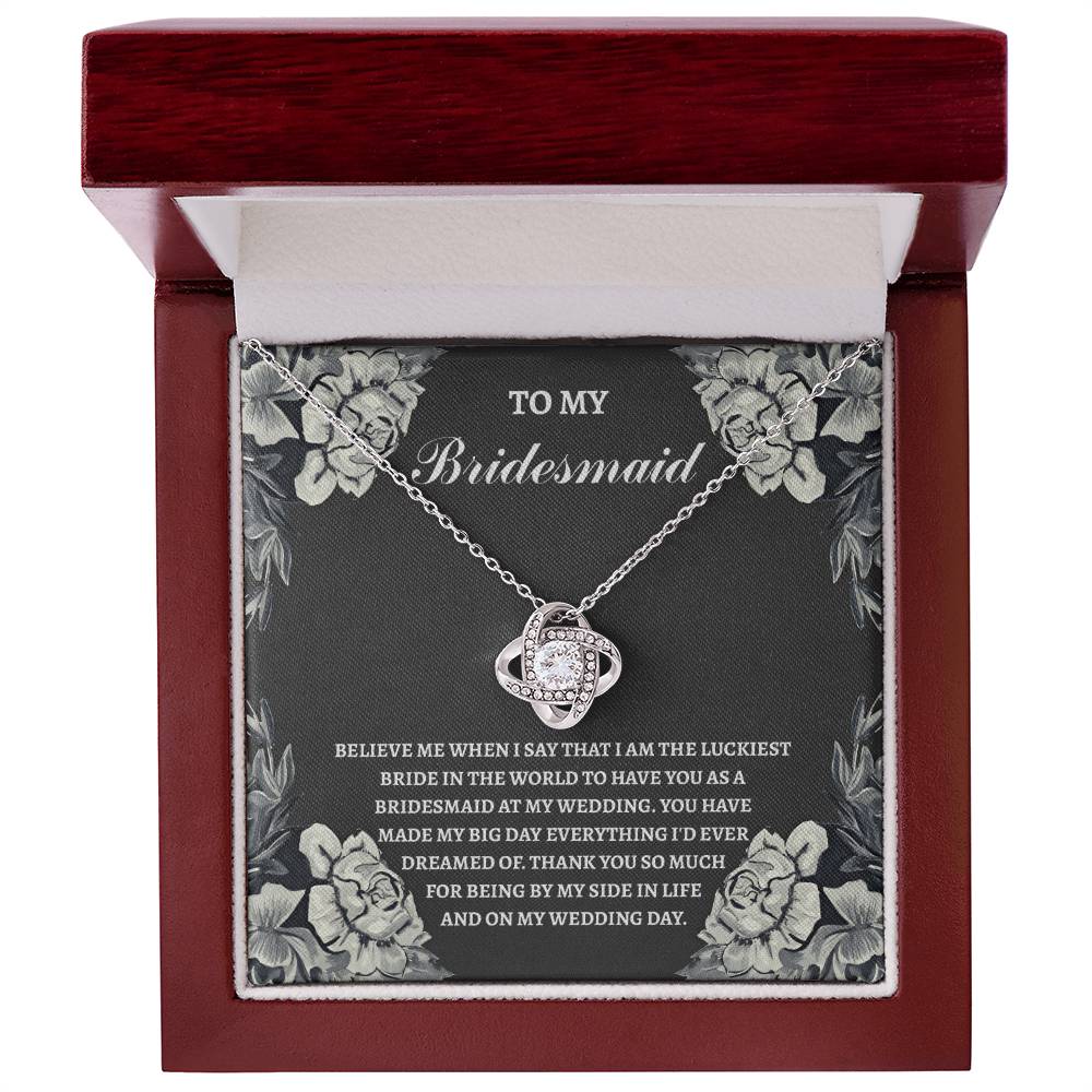 The "To Bridesmaid, The Luckiest Bride - Love Knot Necklace," adorned with cubic zirconia crystals, is elegantly presented on a gift box. The box comes with a heartfelt message expressing gratitude to a bridesmaid. Floral illustrations gracefully decorate the border, adding a charming touch to this piece available in either white gold or yellow gold finish.