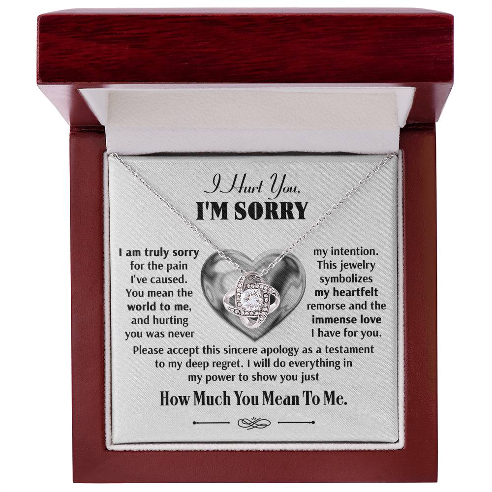 A Sorry-Sincere Apology - Love Knot Necklace displayed in an open jewelry box. The text inside reads an apology message expressing regret and love, with phrases like "I hurt you" and "I'm sorry" alongside sparkling premium cubic zirconia crystals.