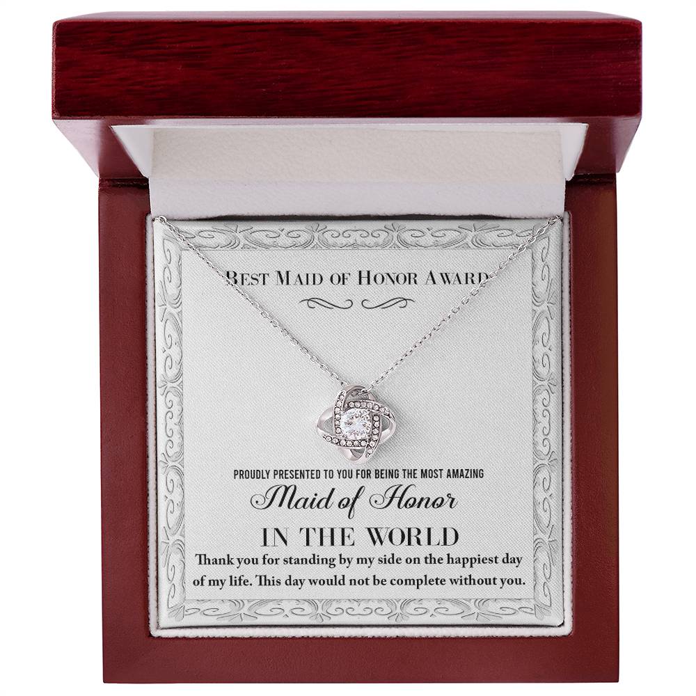 A "To Maid of Honor, Of My Life - Love Knot Necklace," crafted in 14k white gold with a heart-shaped pendant adorned with cubic zirconia crystals, sits elegantly in a jewelry box. The text reads, "Best Maid of Honor Award. Proudly presented to you for being the most amazing Maid of Honor in the world.