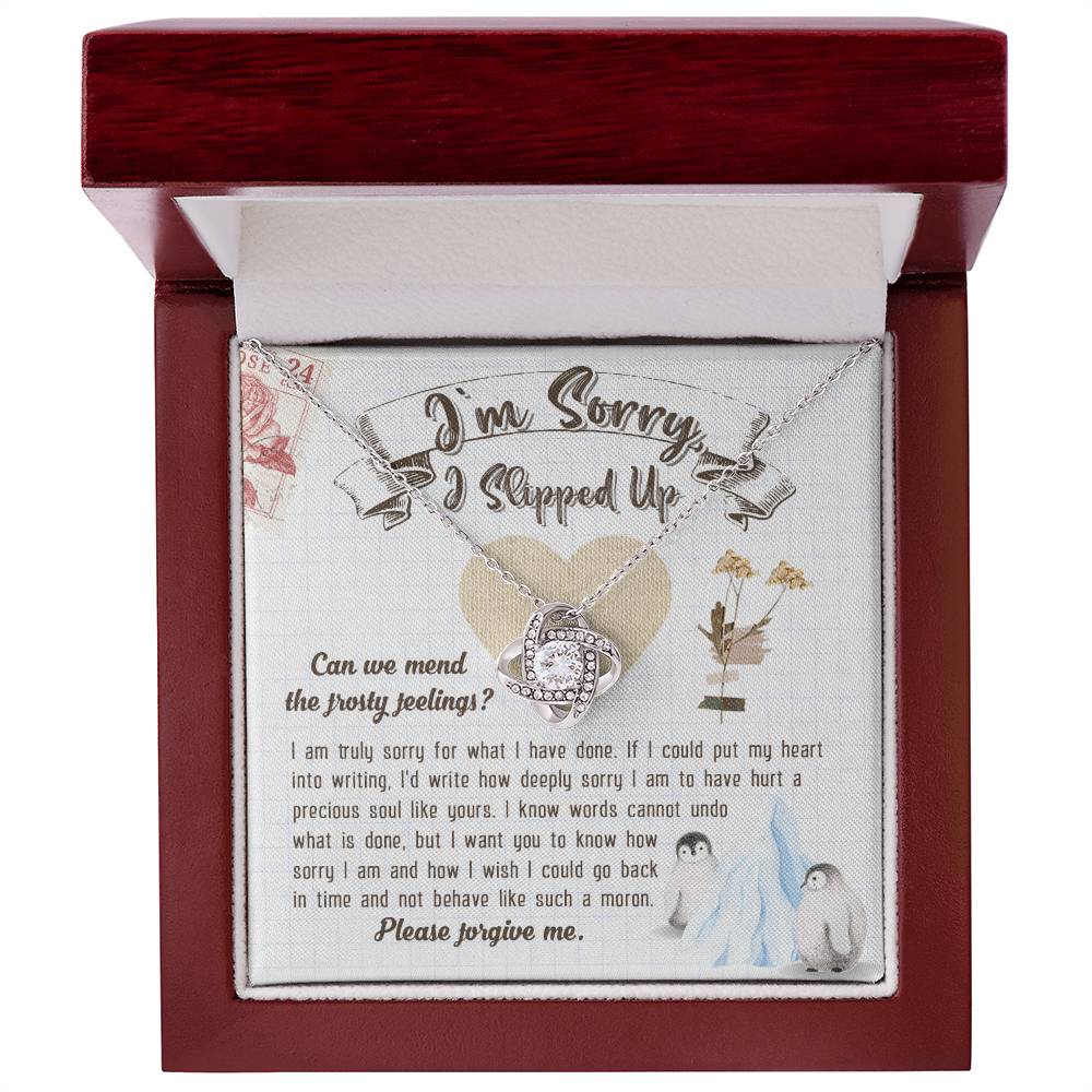 The Sorry-Precious Soul - Love Knot Necklace, featuring an intertwined heart pendant adorned with cubic zirconia crystals, is beautifully displayed in a box. The box comes with an apology note and charming illustrations of flowers, mail stamps, and penguins on a grid-patterned background.