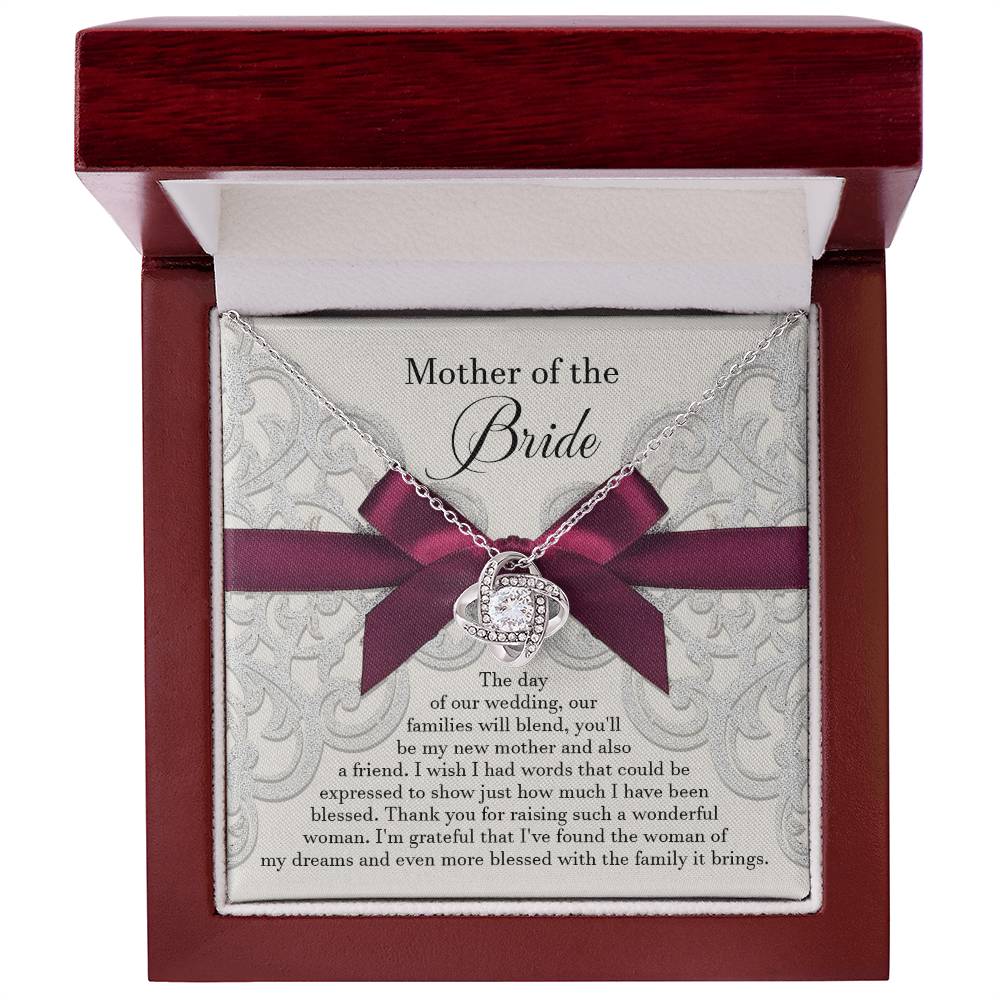 A To Bride's Mom, My New Mother - Love Knot Necklace with a heart pendant, crafted from 14k white gold and adorned with cubic zirconia crystals, is displayed on a decorative card with a maroon ribbon. The card reads, "Mother of the Bride" and contains a heartfelt message of gratitude.