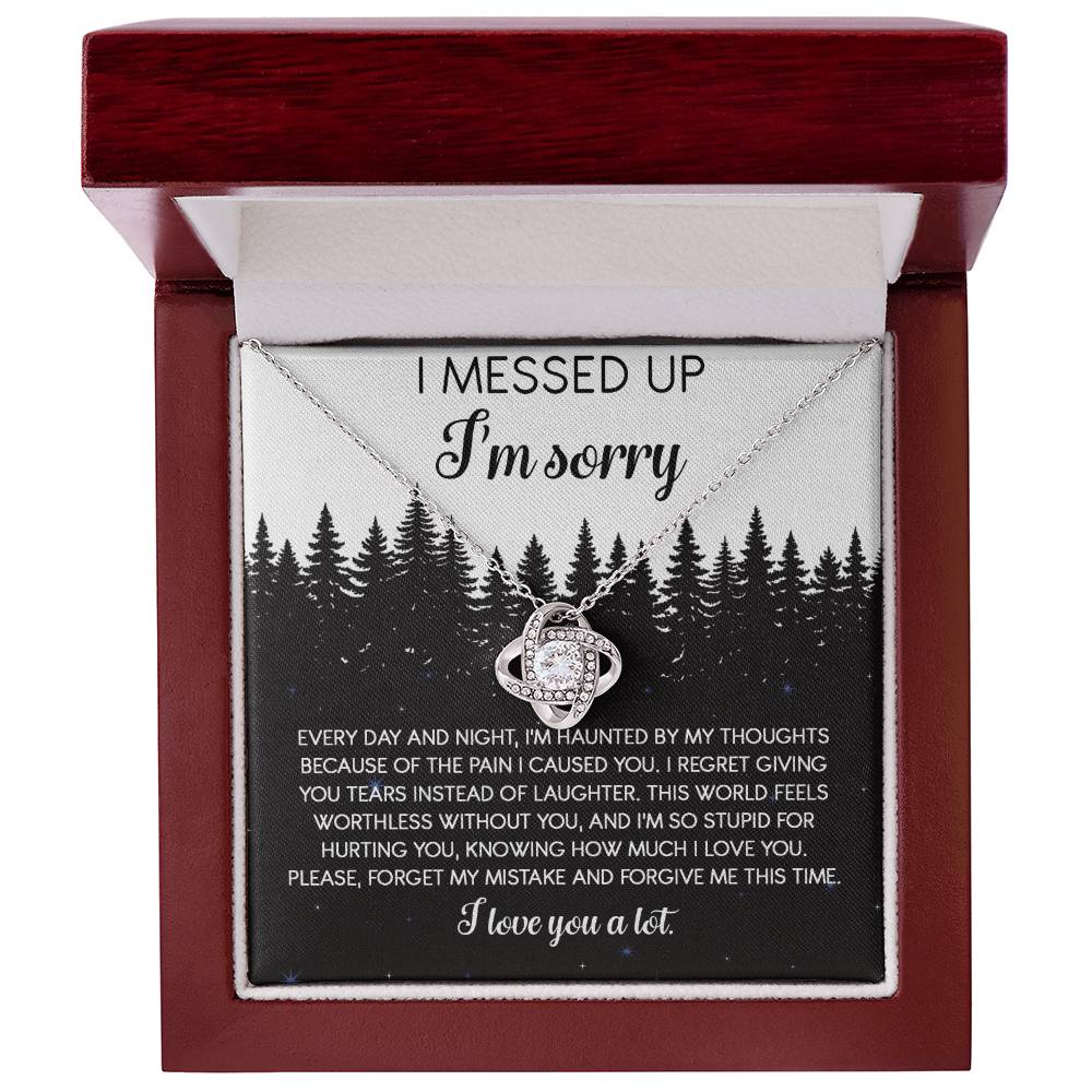 The Sorry-By My Thoughts - Love Knot Necklace, featuring a heart-shaped pendant adorned with cubic zirconia crystals, is displayed on a card with an apology message. The card reads: "I messed up. I'm sorry. Every day and night, I'm haunted...I love you a lot," set against a serene forest background.