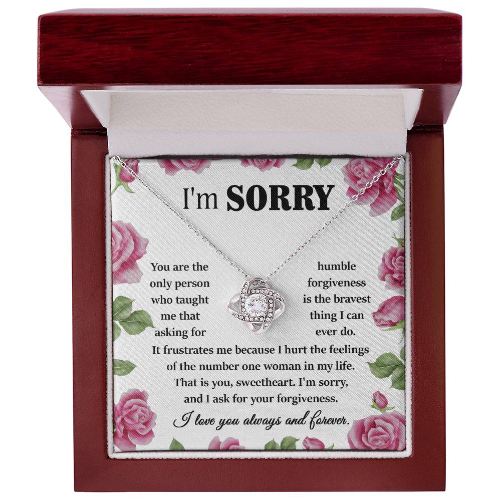 The "Sorry-Your Forgiveness - Love Knot Necklace" features a heart-shaped pendant inside an open red velvet box, adorned with floral decorations and an apology message. This elegant necklace is available in either white gold or yellow gold finishes and sparkles with cubic zirconia crystals.