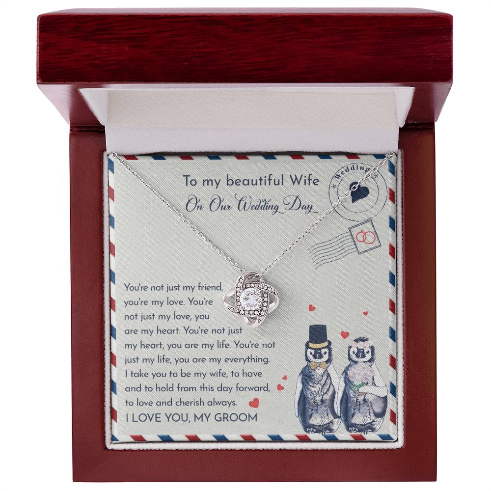 Close-up of a To Wife Wedding, Love And Cherish - Love Knot Necklace with cubic zirconia crystals on a card. The 14k white gold pendant is heart-shaped, and the card has a message for a wife on the wedding day. Two penguins, dressed as a bride and groom, are illustrated at the bottom.