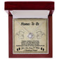 The silver Love Knot Necklace, "Mama To Be-Enjoy The Ride," with a heart and final stone pendant is elegantly displayed in a red jewelry box. The liner, adorned with cherubs and decorative text, features a "Mama-To-Be" message, offering the perfect personalized gift for welcoming parenthood.