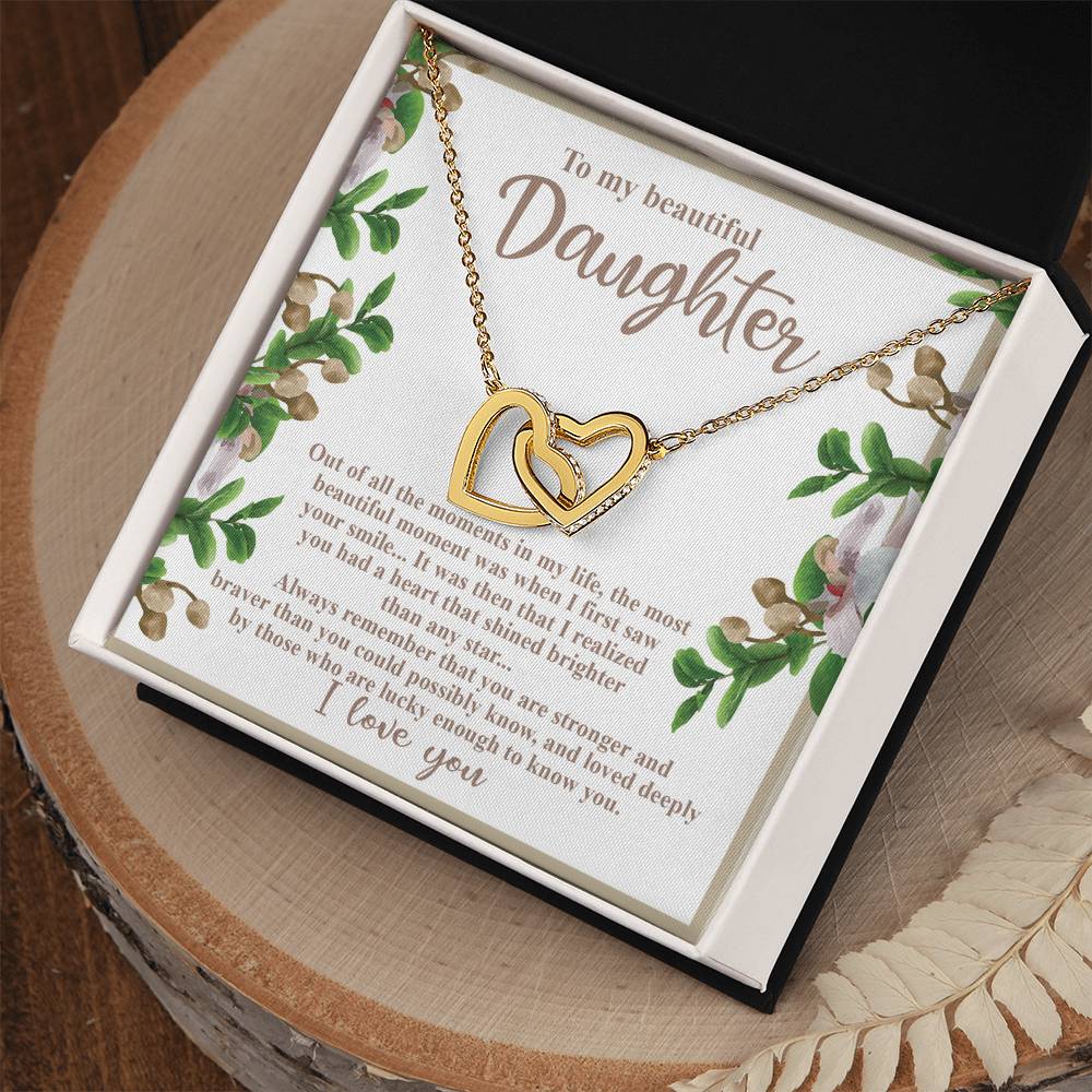 A stunning Daughter-First Smile - Interlocking Hearts Necklace with a rose gold finish rests in a black box, featuring an affectionate message for a daughter. Adorned with floral designs, the card expresses love and admiration, set among white feathers on a wooden surface.