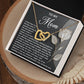The Mom-Guiding Light Interlocking Hearts Necklace, featuring a rose gold finish and sparkling cubic zirconia crystals, sits on a "To My Mom" card with a heartfelt message in a gift box. The box is elegantly displayed on a wooden surface with a beige decorative cloth.