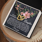 The Daughter-Love Sight necklace, featuring a gold interlocking hearts design with a rose gold finish and floral motifs, is beautifully showcased in a white box. The background includes pink flowers, green leaves, and a loving daughter-oriented message.
