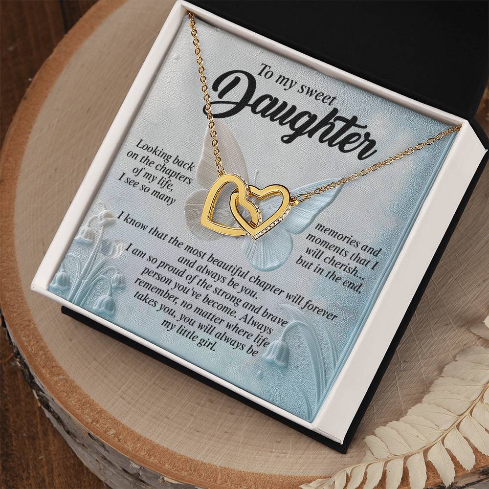 The Daughter-Beautiful Chapter - Interlocking Hearts Necklace, made of gold with cubic zirconia crystals, rests in an open black box. Inside the lid is a heartfelt message to a daughter framed by lily of the valley blooms on blue. The box is elegantly placed on a wooden surface next to a decorative leaf.