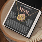 The Mom-More To Me 2 necklace, featuring interlocking hearts with zirconia crystals, is beautifully showcased in a jewelry box with floral designs and heartfelt text, expressing love for a mother. The box sits gracefully on a wooden surface.