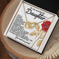 A heartfelt message for a daughter accompanies the Daughter-Journey Challenges - Interlocking Hearts Necklace, featuring cubic zirconia crystals and a red rose illustration. The open rose gold-finished box elegantly rests on a wooden surface.
