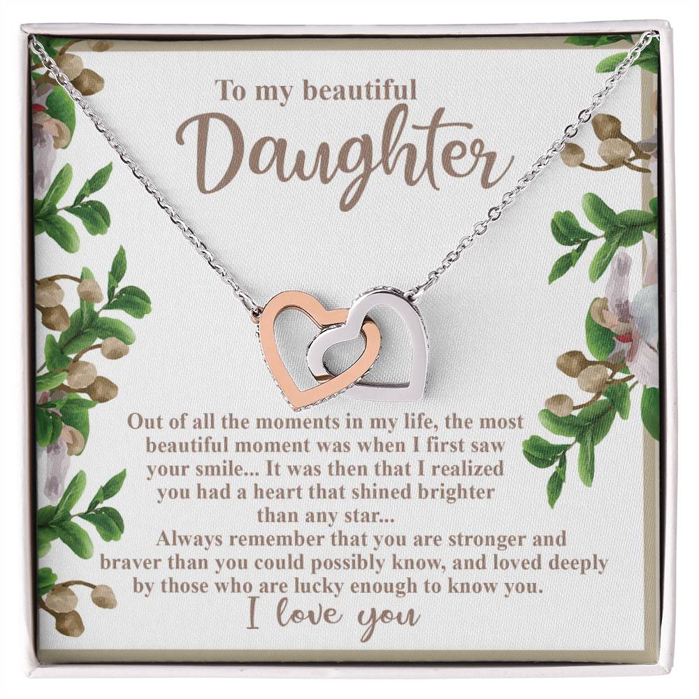 The Daughter-First Smile - Interlocking Hearts Necklace is crafted with surgical steel and a rose gold finish, featuring cubic zirconia crystals. It comes on a decorated card with a heartfelt message to a daughter, surrounded by beautiful floral artwork.