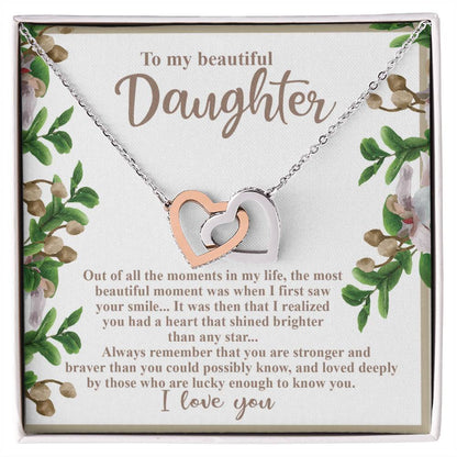 The Daughter-First Smile - Interlocking Hearts Necklace is crafted with surgical steel and a rose gold finish, featuring cubic zirconia crystals. It comes on a decorated card with a heartfelt message to a daughter, surrounded by beautiful floral artwork.