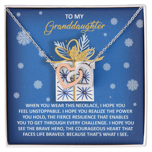 This gift box, ideal for a granddaughter, includes the Granddaughter-Brave Hero - Interlocking Hearts Necklace embellished with cubic zirconia crystals. The silver and gold heart pendant is elegantly displayed against a blue background featuring delicate snowflake patterns, enhancing the enchanting essence of the heartfelt message.