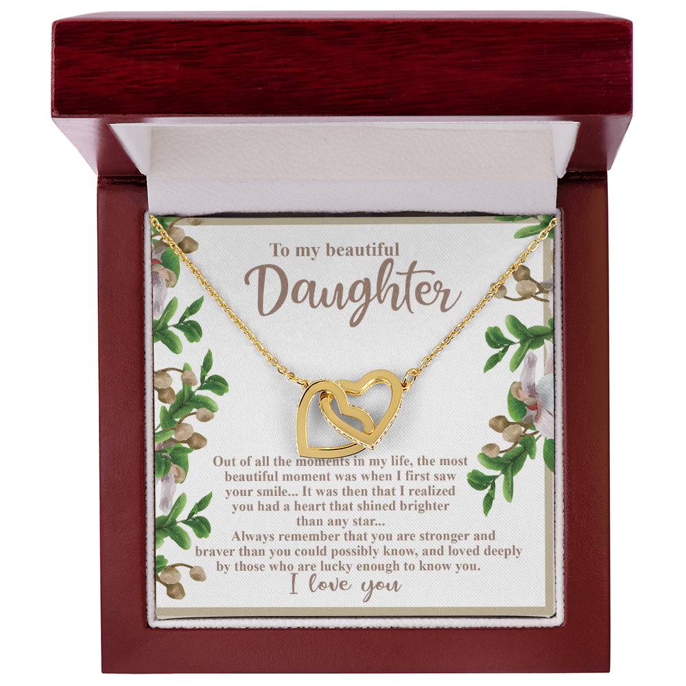The Daughter-First Smile - Interlocking Hearts Necklace, with surgical steel and rose gold, sits in a red jewelry box. The inner lid features a heartfelt message to a daughter, framed by floral designs.