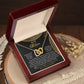 The Mom-Best Friend - Interlocking Hearts Necklace, with a rose gold finish, rests in a red velvet box featuring "Mom" on the background. Dried flowers and fern leaves accentuate the setting on a wooden surface, while cubic zirconia crystals gleam softly.