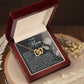 A Mom-Guiding Light - Interlocking Hearts Necklace with a rose gold finish rests in a dark red jewelry box. A "To My Mom" card, featuring a rose design and heartfelt message, stands behind. The box sits on a wooden surface adorned with decorative dried flowers for elegance.