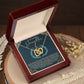 The Daughter-Little Princess Interlocking Hearts Necklace features a rose gold finish with sparkling cubic zirconia crystals, elegantly displayed in an open wooden box on a round surface adorned with dried flowers. Inside, a card reads, "To my beautiful daughter.