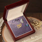 The Mom-Shining Star - Interlocking Hearts Necklace in rose gold, featuring a heart pendant, lies in an open wooden box. Inside, it reads "Mom," surrounded by butterfly illustrations and sparkling cubic zirconia. The box on a wooden surface is scattered with dried flowers.