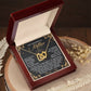 The Mom-God's Masterpiece Interlocking Hearts Necklace, featuring a rose gold finish and sparkling cubic zirconia crystals, is elegantly displayed in an open red velvet-lined jewelry box with a heartfelt message for mothers. The setting includes a wooden background adorned with delicate dried flowers.
