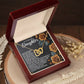 The Daughter-Loved Enough - Interlocking Hearts Necklace with cubic zirconia crystals, nestled in a wooden jewelry box, gracefully drapes over a rose-designed card featuring a heartfelt message. Dried flowers on the wood surface enhance its charm.