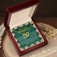 The "Daughter-Your Journey - Interlocking Hearts Necklace" in a red jewelry box features a note saying: "To My Beautiful Daughter." Made of surgical steel and rose gold, it shines with cubic zirconia crystals, displayed open on a wooden surface surrounded by dried flowers.