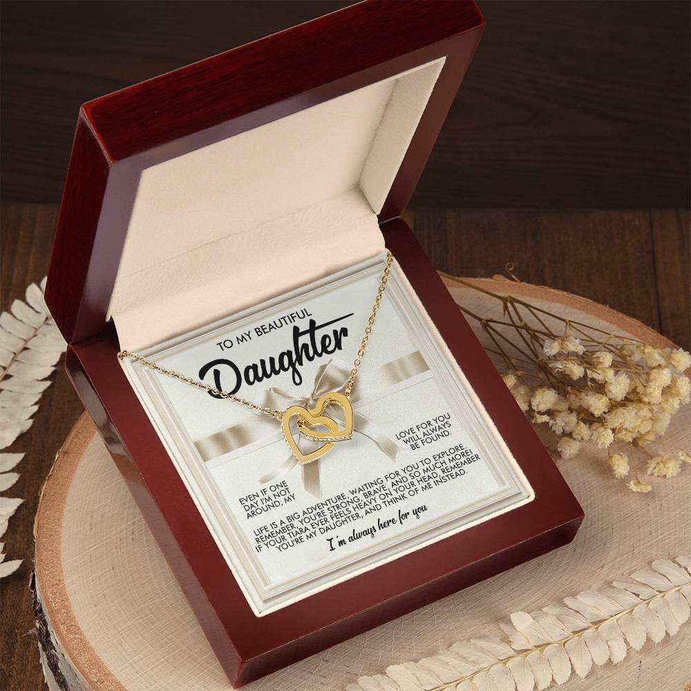 The Daughter-Heavy Tiara - Interlocking Hearts Necklace, a gold heart-shaped piece with a small gem, sits elegantly in an open red jewelry box. The rose gold finish and nearby dried flowers enhance its charm. A heartfelt message inside the lid makes this necklace truly special.